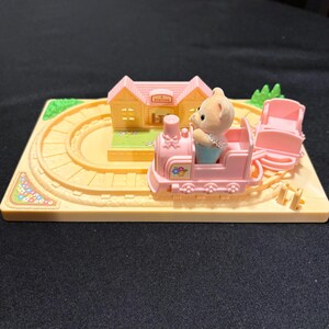 Pode incluir: Um conjunto de comboio de brinquedo em miniatura com um comboio rosa, uma figura de urso, uma esta&ccedil;&atilde;o e uma pista. O comboio tem detalhes florais e puxa uma carruagem rosa. A esta&ccedil;&atilde;o &eacute; amarela com um telhado rosa.