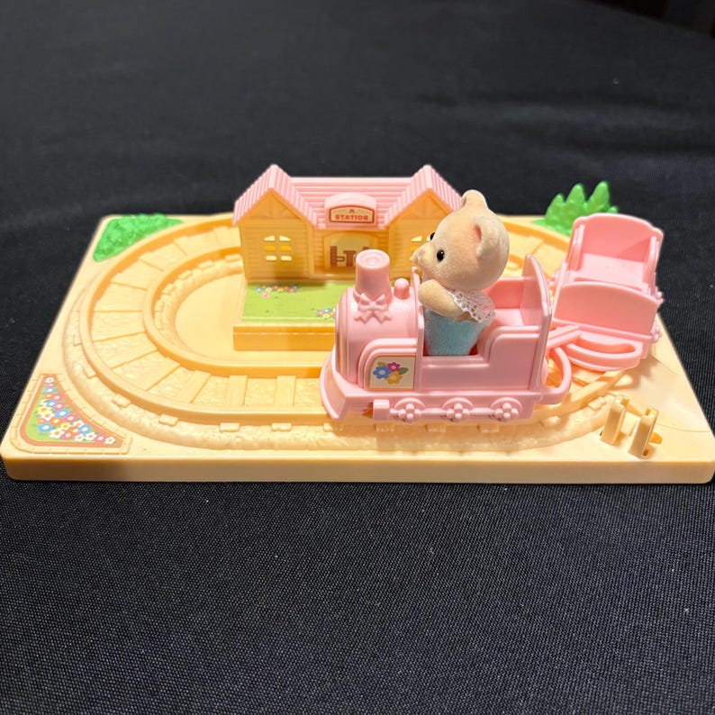 Pode incluir: Um conjunto de comboio de brinquedo em miniatura com um comboio rosa, uma figura de urso, uma esta&ccedil;&atilde;o e uma pista. O comboio tem detalhes florais e puxa uma carruagem rosa. A esta&ccedil;&atilde;o &eacute; amarela com um telhado rosa.