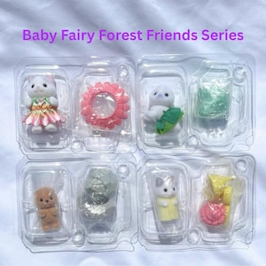 May include: Baby Fairy Forest Friends Series set featuring four small flocked animal figurines in clear plastic packaging. The set includes a white cat in a floral dress, a white seal with a green leaf, a brown bear, and a white cat in yellow.
