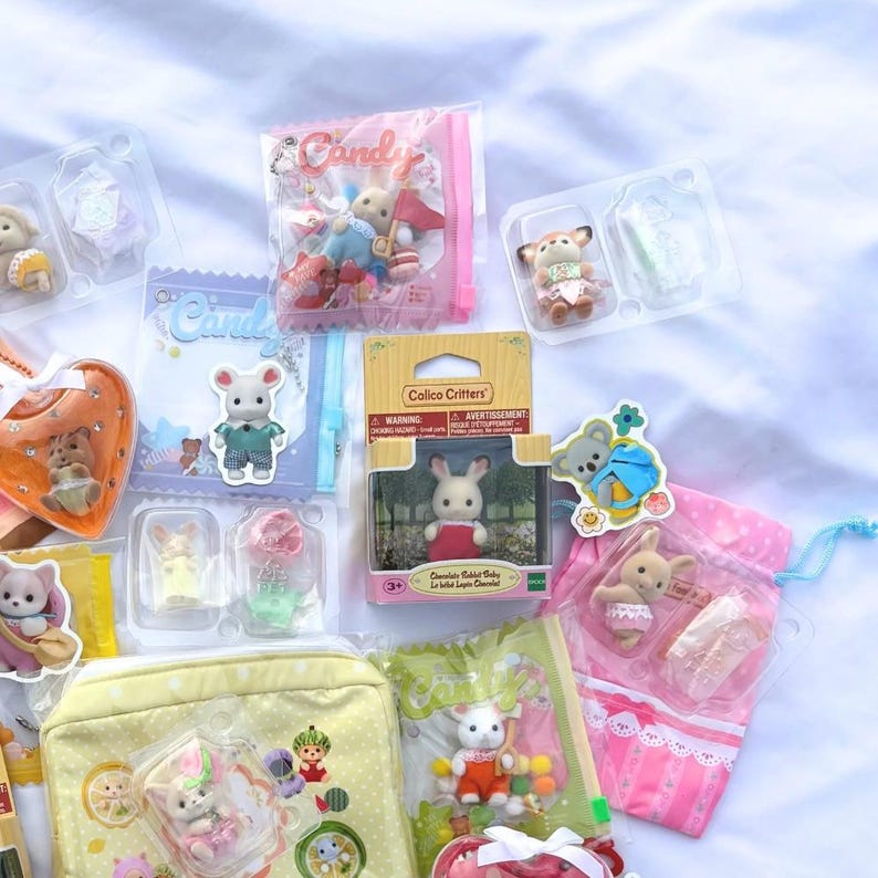 May include: A collection of Calico Critters toys, including animal figures, accessories, and packaging. The toys are in various colors, including pink, yellow, and white. Some are packaged in clear plastic, while others are in bags or boxes.