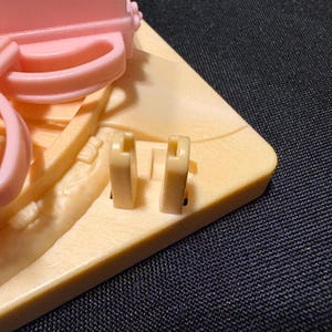 Pode incluir: Close-up de um brinquedo de pl&aacute;stico rosa claro e bege. O brinquedo apresenta uma pega, dois ganchos bege e uma base bege. O fundo &eacute; um tecido azul escuro.