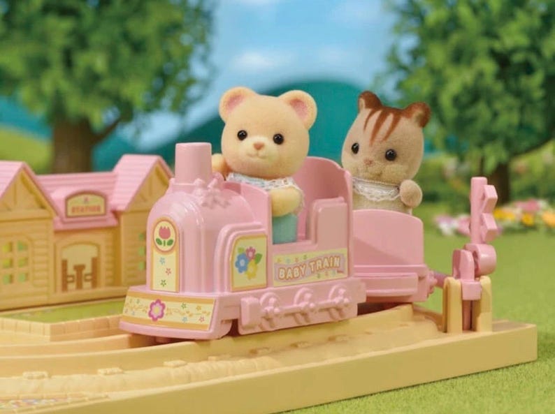 Pode incluir: Um comboio de brinquedo rosa com um urso e um gato como passageiros. O comboio tem a inscri&ccedil;&atilde;o "Baby Train" na lateral. O comboio est&aacute; sobre uma via de cor bege claro com uma esta&ccedil;&atilde;o amarela ao fundo. A cena &eacute; ao ar livre.