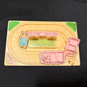 Pode incluir: Um conjunto de comboio de brinquedo em miniatura com uma locomotiva rosa claro e dois vag&otilde;es sobre uma via bege. Uma pequena figura de urso com macac&atilde;o azul est&aacute; perto de um edif&iacute;cio rosa. O conjunto inclui detalhes verdes e florais.