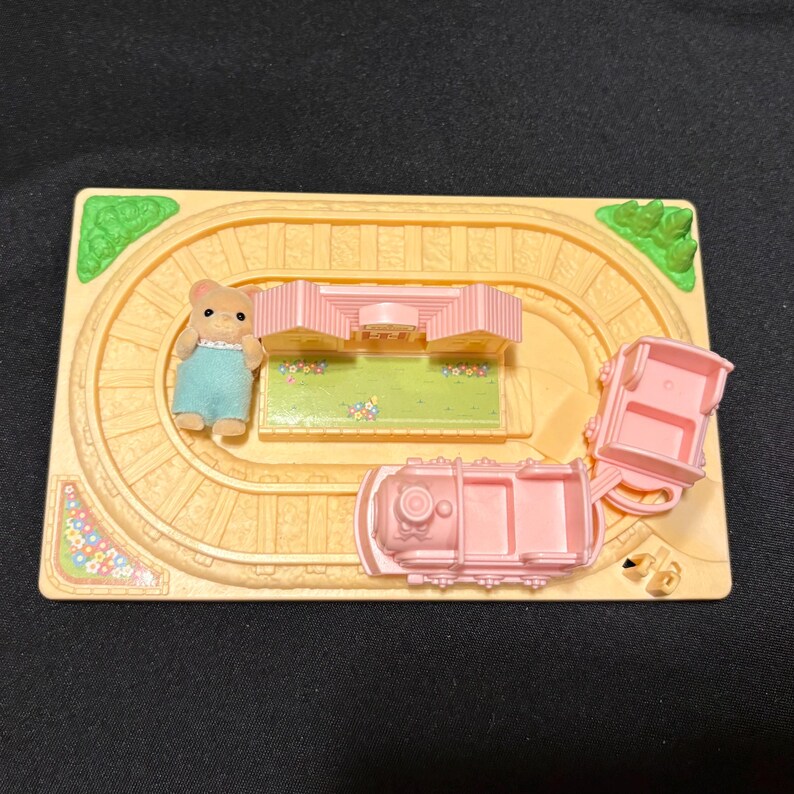 Pode incluir: Um conjunto de comboio de brinquedo em miniatura com uma locomotiva rosa claro e dois vag&otilde;es sobre uma via bege. Uma pequena figura de urso com macac&atilde;o azul est&aacute; perto de um edif&iacute;cio rosa. O conjunto inclui detalhes verdes e florais.