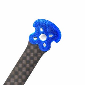 Op de afbeelding: Een close-up van een blauw, 3D-geprint onderdeel met een unieke vorm, met meerdere gaten. Het is bevestigd aan een carbon fiber arm met een dambordpatroon. De achtergrond is effen wit.