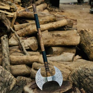 Może przedstawiać: Topór dwustronny z głowicą z metalu w kolorze srebrnym, ozdobioną misternymi grawerowanymi wzorami węzłów. Długi drewniany trzonek jest udekorowany rzeźbionymi geometrycznymi motywami i owinięty czarnym sznurkiem w górnej i środkowej części.
