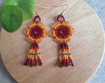 Puede incluir: Un par de pendientes de cuentas con un diseño de flor en tonos de granate, amarillo y dorado. Los pendientes tienen una franja tipo borla.