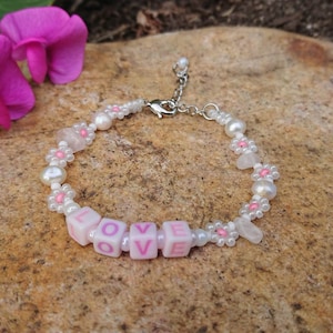 Könnte beinhalten: Ein weißes Perlenarmband mit rosa Blumenakzenten und rosa Buchstabenperlen, die "LOVE" buchstabieren.