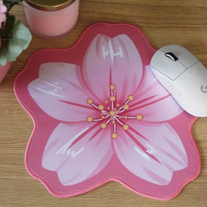 May include: A flower-shaped mouse mat featuring a large pink cherry blossom design with five petals, a central cluster of yellow stamens, and white highlights. The mouse mat is positioned on a wooden surface next to a white wireless computer mouse.