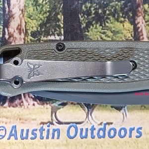 May include: A green pocket knife with a black handle and a silver clip. The knife has a textured handle and a black blade. The knife is made by the company "ADCS".