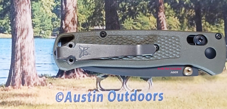 May include: A green pocket knife with a black handle and a silver clip. The knife has a textured handle and a black blade. The knife is made by the company "ADCS".