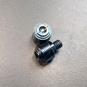 Könnte beinhalten: Zwei kleine Metallkomponenten mit dunkler, irisierender Oberfläche. Eines ist zylindrisch mit konzentrischen Ringen, das andere hat eine Gewinde-Sechskantform. Die Objekte sind wahrscheinlich Hardware oder Befestigungselemente.