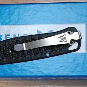 May include: A black pocket knife with a silver clip and a butterfly logo. The knife is closed and has a textured handle. The knife is on a blue box with the words "BUGOUT" and "MANUAL OPEN" printed on it.