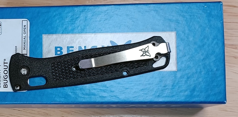 May include: A black pocket knife with a silver clip and a butterfly logo. The knife is closed and has a textured handle. The knife is on a blue box with the words "BUGOUT" and "MANUAL OPEN" printed on it.