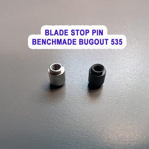 May include: Two blade stop pins for a Benchmade Bugout 535 knife. One pin is silver-colored, and the other is black. The pins are cylindrical with a threaded hole at the top. Text on the image reads "BLADE STOP PIN BENCHMADE BUGOUT 535."
