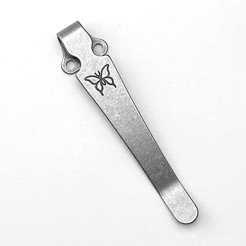 May include: A silver pocket clip with a butterfly design. The clip has two holes on the top and a rounded end.