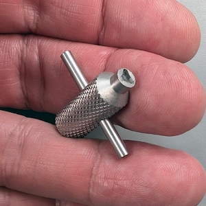 May include: A small, silver metal tool with a knurled grip and a hexagonal socket. Two cylindrical bars extend from the tool's body, forming a cross shape. The tool is designed for tightening or loosening screws or bolts.