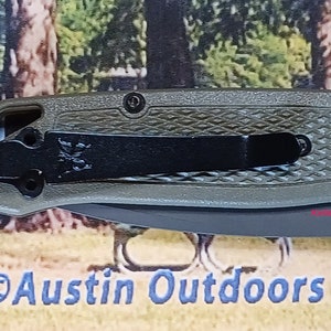 May include: A green pocket knife with a black clip and a textured handle. The knife has a black blade and a silver pocket clip. The knife is made by the company "ADCS".