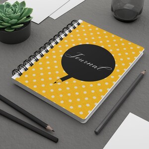 May include: A yellow journal with white polka dots and a black circle with the word "Journal" written in white cursive. The journal is lying open on a grey surface with two black pencils and a white envelope.