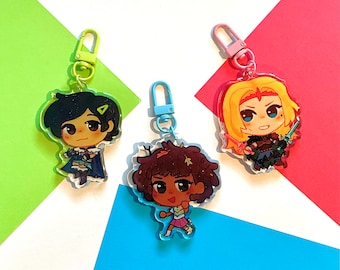 May include: Three colorful keychains featuring cartoon characters. The first keychain features a girl with dark blue hair and a green outfit. The second keychain features a girl with brown hair and a pink outfit. The third keychain features a girl with blonde hair and a black and white outfit.
