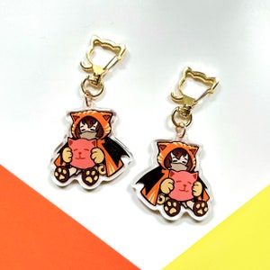 Op de afbeelding: Twee oranje en bruine sleutelhangers met een kattenmotief en gouden sluitingen. Elke sleutelhanger heeft een cartoonfiguur dat een capuchon draagt en een roze pluche kat vasthoudt.