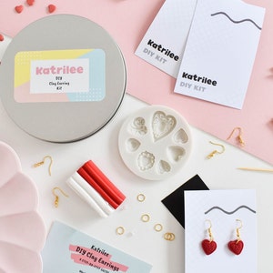May include: A craft kit for making clay earrings, featuring a silver tin, clay, earring hooks, and a heart-shaped mould. The kit includes instructions and finished red heart earrings. The tin is labelled "Katrilee DIY Clay Earring Kit."