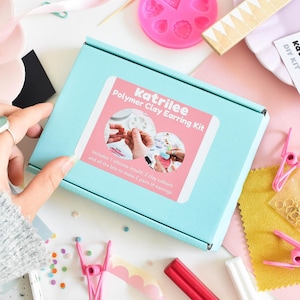 May include: A light blue box labeled "Katrilee Polymer Clay Earring Kit" with a pink and white sticker. The kit includes a silicone mold, clay colors, and earring-making supplies. Other items include pink clips, gold rings, and a pink heart-shaped mold.
