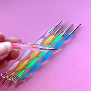May include: A collection of nail art dotting tools with clear, spiral-patterned handles in various colors, including pink, yellow, green, and blue. Each tool has a metal tip for creating precise designs. One tool is held by a hand.