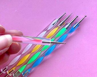 Può includere: Un set di strumenti per il dotting nail art con manici trasparenti a spirale in vari colori, tra cui rosa, giallo, verde e blu. Ogni strumento ha una punta metallica per creare disegni precisi. Uno strumento è tenuto da una mano.