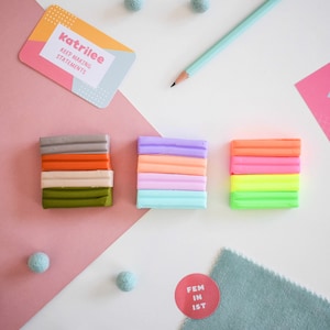 May include: A flat lay featuring colorful clay bars in stacks, a pencil, a business card with the text "katrilee KEEP MAKING STATEMENTS", felt balls, and a sticker with the text "FEM IN IST". The background is a mix of white, pink, and teal.