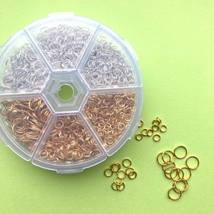 May include: A clear plastic organiser containing silver and gold jump rings. Several jump rings are scattered outside the organiser on a light green surface. The jump rings are in various sizes, ideal for jewellery making and crafts.