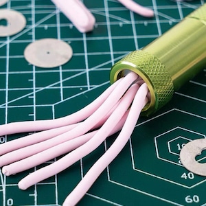 Puede incluir: Primer plano de una herramienta de extrusión de metal verde con hebras de arcilla rosa saliendo. La herramienta está sobre una esterilla de corte verde con piezas metálicas circulares y otros trozos de arcilla. La escena está bien iluminada y enfocada en el proceso de elaboración.