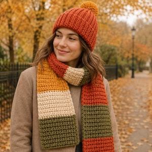 Op de afbeelding: Een geribbelde gebreide muts in roestoranje met een bijpassende pompon, gecombineerd met een lange, kleurgeblokte sjaal met horizontale strepen in roestoranje, mosterdgeel, crème en olijfgroen, gedragen over een beige jas.