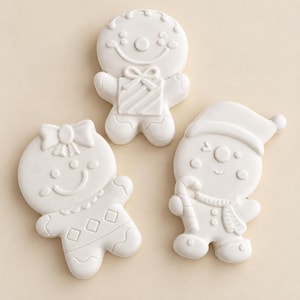 May include: Three white, unpainted ceramic gingerbread-style figures. One figure features a bow on its head, another holds a gift, and the third wears a hat and scarf while holding a candy cane. Each figure has embossed facial and clothing details.