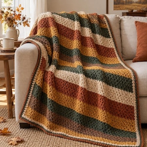 Puede incluir: Una manta de ganchillo con rayas horizontales en colores alternos crema, amarillo mostaza, verde bosque y naranja quemado. La manta presenta un patrón de puntos texturizado y grueso con un borde acabado en hilo color crema.