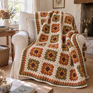May include: A handmade crochet blanket featuring a repeating granny square pattern in a color palette of cream, mustard yellow, burnt orange, olive green, and deep plum. The square motifs are joined together with a cream-colored yarn border.