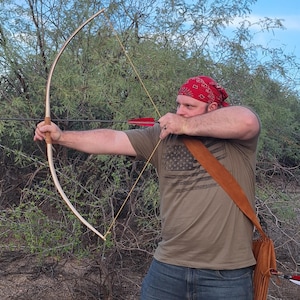 May include: A wooden longbow with a drawn arrow, held by a person wearing a red bandana and olive green t-shirt. The bow has a light wood finish, and the arrow has red fletching. A brown leather quiver is visible.