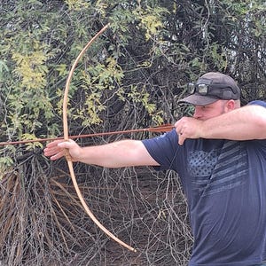 Puede incluir: Un arco largo de madera está tensado, listo para disparar una flecha. El arco es de color marrón claro y la flecha es marrón con un extremo emplumado. La persona lleva una camiseta azul oscuro con un diseño de bandera.