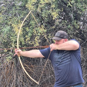 Puede incluir: Una persona tensando un arco y una flecha de madera, lista para disparar. El arco es de madera de color claro y la flecha es marrón. La persona lleva una camiseta azul oscuro con un diseño de bandera y una gorra de béisbol.