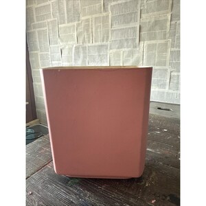 Puede incluir: Un contenedor cuadrado de color rosa polvoriento con una superficie ligeramente texturizada. El contenedor está sobre una mesa de madera oscura. El fondo es una pared cubierta de recortes de periódico.