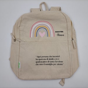 May include: A beige canvas backpack with a rainbow and heart design. The backpack has a zipper closure and adjustable straps. The text "MAESTRA Chiara" is printed on the front of the backpack. The quote "Ogni persona che incontri ha qualcosa di simile a te e qualcos'altro di tanto favoloso che non ti somiglia per niente" is printed on the bottom of the backpack.