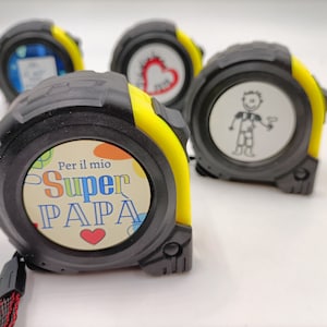 Può includere: Un metro a nastro giallo e nero con la scritta "Per il mio Super PAPA" e un cuore rosso sul davanti.