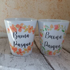 Può includere: Due tazze in ceramica bianca con un design floreale e il testo "Buona Pasqua" in lettere nere. Le tazze sono su una superficie di legno bianco.
