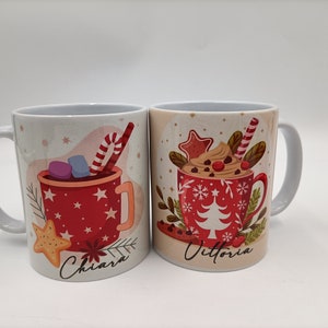 Può includere: Due tazze in ceramica bianca con disegni a tema natalizio rosso e bianco. La tazza a sinistra ha una tazza rossa con marshmallow e una canna di zucchero, e il nome "Chiara" scritto sotto. La tazza a destra ha una tazza rossa con panna montata, gocce di cioccolato e una canna di zucchero, e il nome "Vittoria" scritto sotto.