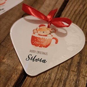 Può includere: Ciondolo natalizio bianco a forma di cuore con un nastro rosso. Il ciondolo presenta una volpe di cartone animato con un cappello di Babbo Natale in una tazza rossa con il testo "Buon Natale" e il nome "Silvia".