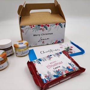 Può includere: Una scatola regalo di Natale in cartone marrone con un design floreale. La scatola ha un manico ed è decorata con le parole "Buon Natale Laura". La scatola contiene tre piccoli vasetti di marmellata, una tavoletta di cioccolato e un biglietto.