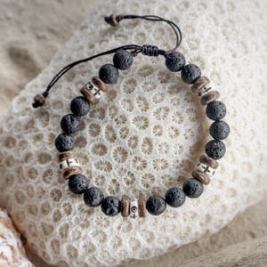 May include: A beaded bracelet featuring black lava stone beads and decorative wooden and bone accents. The bracelet is strung on a dark brown cord and is displayed on a white, textured surface. The bracelet is a stylish accessory.