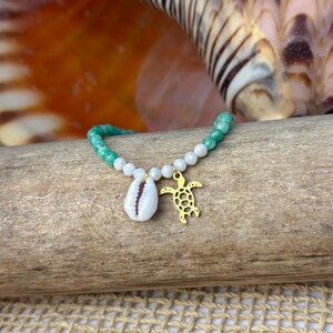 Puede incluir: Una pulsera de cuentas con una concha blanca, un dije de tortuga dorada y cuentas verdes y blancas. La pulsera se exhibe sobre un trozo de madera, con una concha en el fondo.