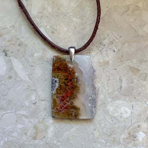 Pode incluir: Um pendente retangular com uma fatia de pedra natural com inclusões em castanho terra, verde musgo e vermelho vibrante, contornado por uma borda branca translúcida e um lado cristalino irregular, suspenso por um colar de cordão de couro castanho escuro.