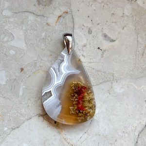 May include: A teardrop-shaped pendant featuring a polished stone with translucent white and gray banding on one side and a warm amber-toned section on the other, containing moss-like green inclusions and a distinct bright red mineral formation.